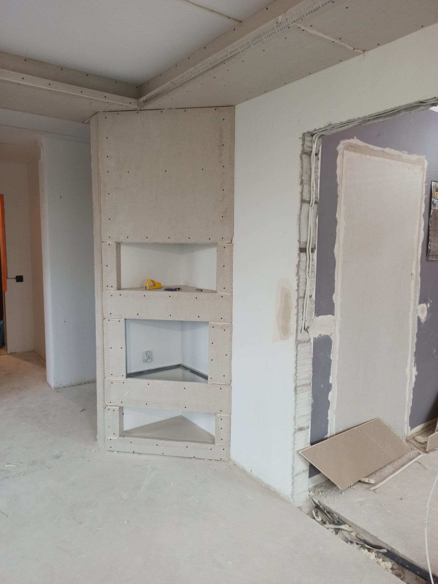 Wnętrze w trakcie remontu: ściana z płyt gipsowo-kartonowych z wnękami, widoczne przewody elektryczne i narzędzia, surowy beton na podłodze.