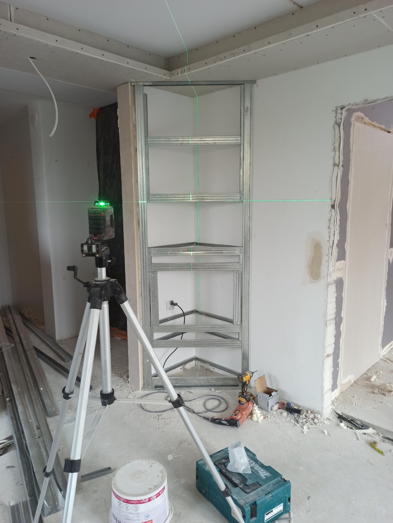 Metalowa konstrukcja narożnej półki w trakcie montażu, wyznaczana laserem krzyżowym, widoczny sprzęt budowlany i narzędzia