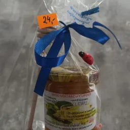 Słoik miodu lipowego z drewnianym nabierakiem, zapakowany w celofan z niebieską wstążką i ozdobną biedronką, prezentowany w dłoni.