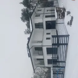 Dwukondygnacyjny dom murowany z białymi ścianami i czarnymi ramami okiennymi, ogrodzony czarnym, metalowym płotem, w zimowej scenerii, z paletami obok.