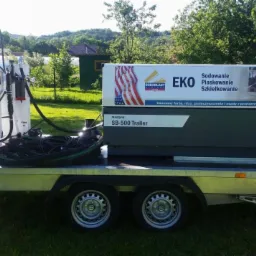 Mobilna maszyna do obróbki strumieniowo-ściernej, typu SB-500 Trailer, z białym zbiornikiem i czarnymi przewodami, umieszczona na przyczepie w plenerze.