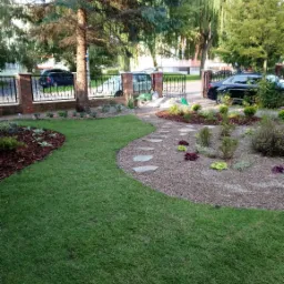 Świeżo założony trawnik z rolki, graniczący z rabatą wysypaną grysem i ścieżką z kamieni, oddzielający ogród od kutej bramy wjazdowej i parkingu.