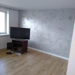 Pokój po odświeżeniu: ściana z dekoracyjną szarą fakturą, drewniana podłoga, telewizor na szafce RTV z widocznymi kablami i otwartymi drzwiczkami.