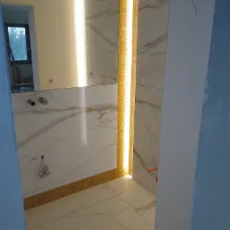 Wnętrze łazienki w trakcie prac wykończeniowych z marmurowymi płytkami, podświetlanymi listwami dekoracyjnymi w kształcie plastra miodu oraz widocznymi elementami instalacji wodnej.