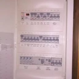 Otwarta skrzynka rozdzielcza z bezpiecznikami i oznaczeniami obwodów, widoczne liczne wyłączniki nadprądowe i różnicowoprądowe, listwy zaciskowe oraz ograniczniki przepięć.