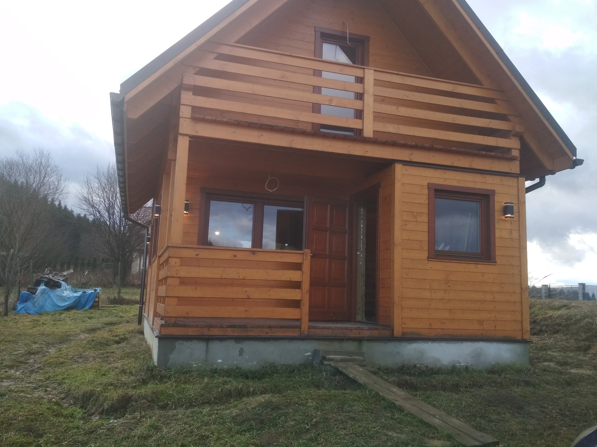 Fasada nowego, drewnianego domu z balkonem, oświetlona lampami zewnętrznymi, z tymczasowym wejściem z desek, na tle pochmurnego nieba i drzew bez liści.