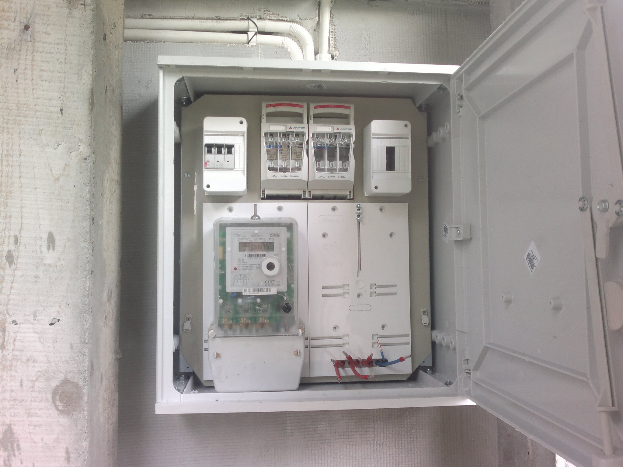 Otwarta skrzynka elektryczna z zamontowanym licznikiem i bezpiecznikami, widoczna na tle surowego betonowego muru.