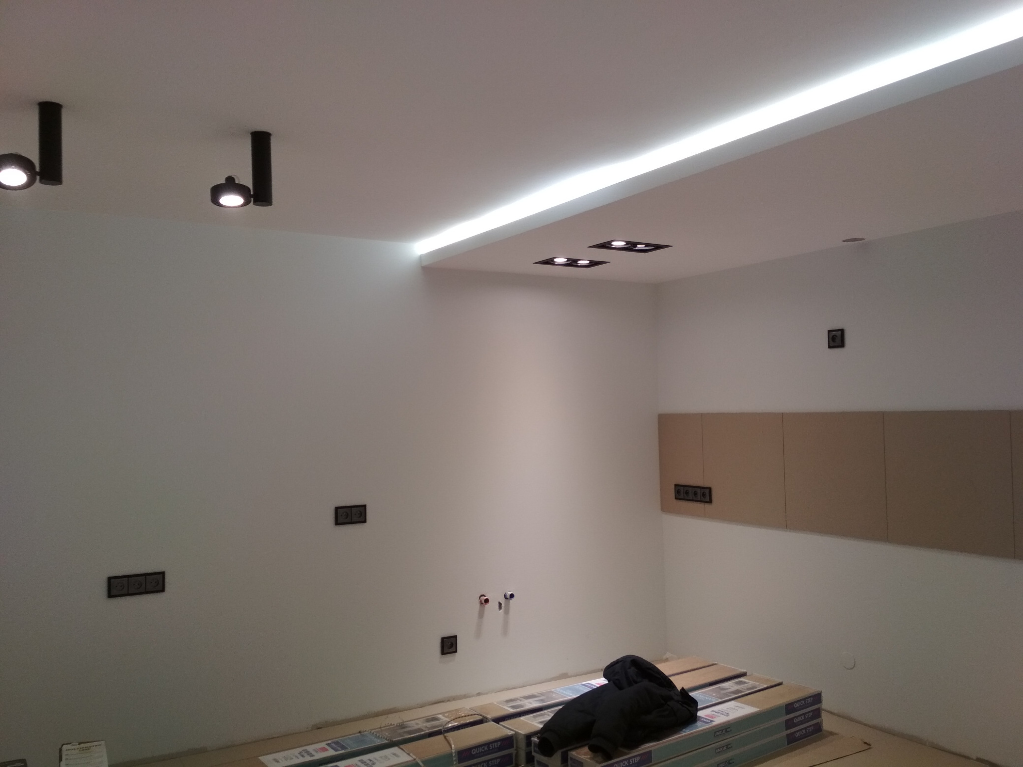 Wnętrze w trakcie wykańczania: zamontowane oświetlenie sufitowe LED liniowe i punktowe, widoczne puszki elektryczne, rury instalacji wodnej i ułożone paczki paneli podłogowych.