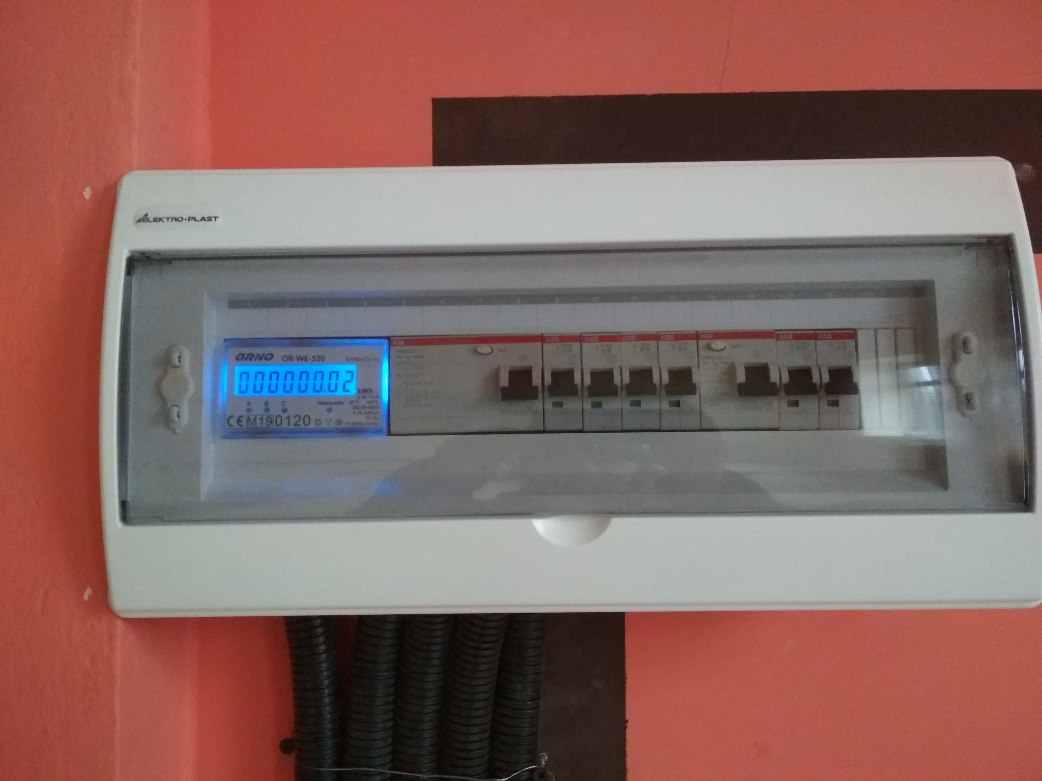 Zamknięta skrzynka rozdzielcza z widocznym licznikiem energii elektrycznej ORNO OR-WE-520 na pomarańczowej ścianie, z czarnymi rurami karbowanymi pod spodem.