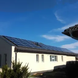 Panele fotowoltaiczne na dachu jednopiętrowego domu z jasną elewacją, widok z dołu w słoneczny dzień.