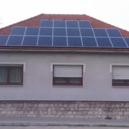 Dom jednorodzinny z panelami słonecznymi na dachu i częściowo opuszczonymi roletami zewnętrznymi w oknach.