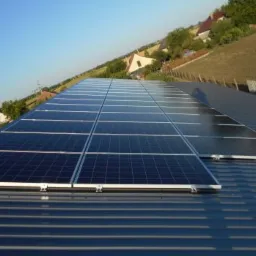 Panel słoneczny zainstalowany na metalowym dachu z widokiem na wiejski krajobraz i błękitne niebo.