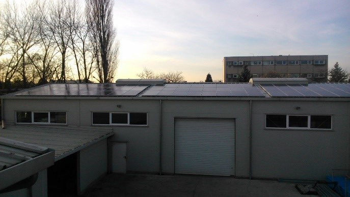 Instalacja paneli słonecznych na dachu magazynu z widocznym budynkiem biurowym w tle, ujęcie z lekkiej perspektywy o zmierzchu.