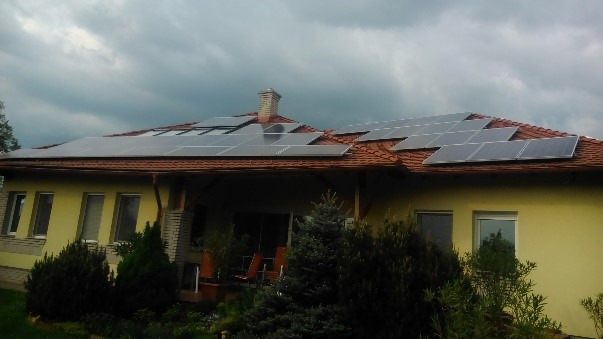 Dom jednorodzinny z zainstalowanymi panelami fotowoltaicznymi na dachu pokrytym czerwoną dachówką, widok z poziomu ogrodu z zielenią i krzewami.