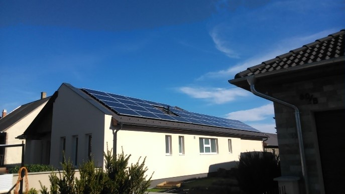 Panele fotowoltaiczne na dachu jednopiętrowego domu z jasną elewacją, widok z dołu w słoneczny dzień.