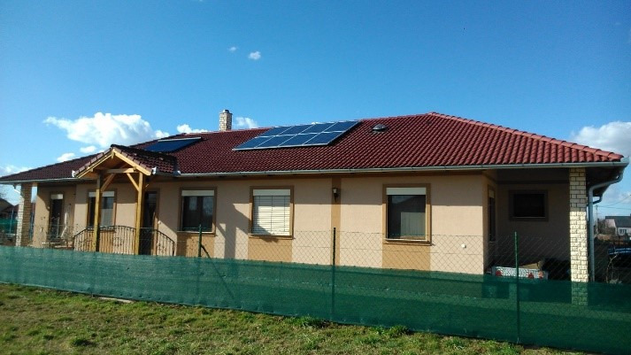 Jednopiętrowy dom z beżową elewacją, czerwoną dachówką i panelami słonecznymi, widok zza zielonego ogrodzenia.