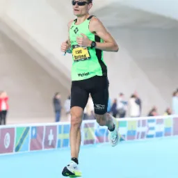 Mężczyzna w okularach przeciwsłonecznych i sportowym stroju biegnie podczas maratonu, widoczny numer startowy 1360, na nadgarstku zegarek sportowy, w tle publiczność i elementy infrastruktury.