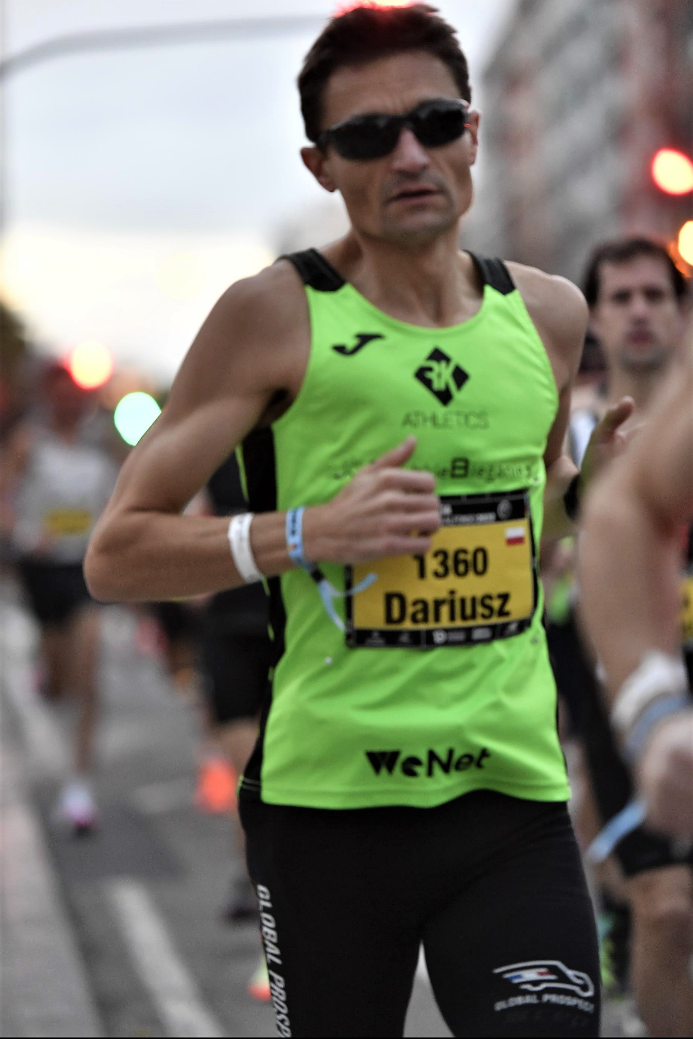 Mężczyzna w okularach przeciwsłonecznych biegnie w maratonie, ubrany w neonowo zielony top z numerem 1360 i czarne legginsy.