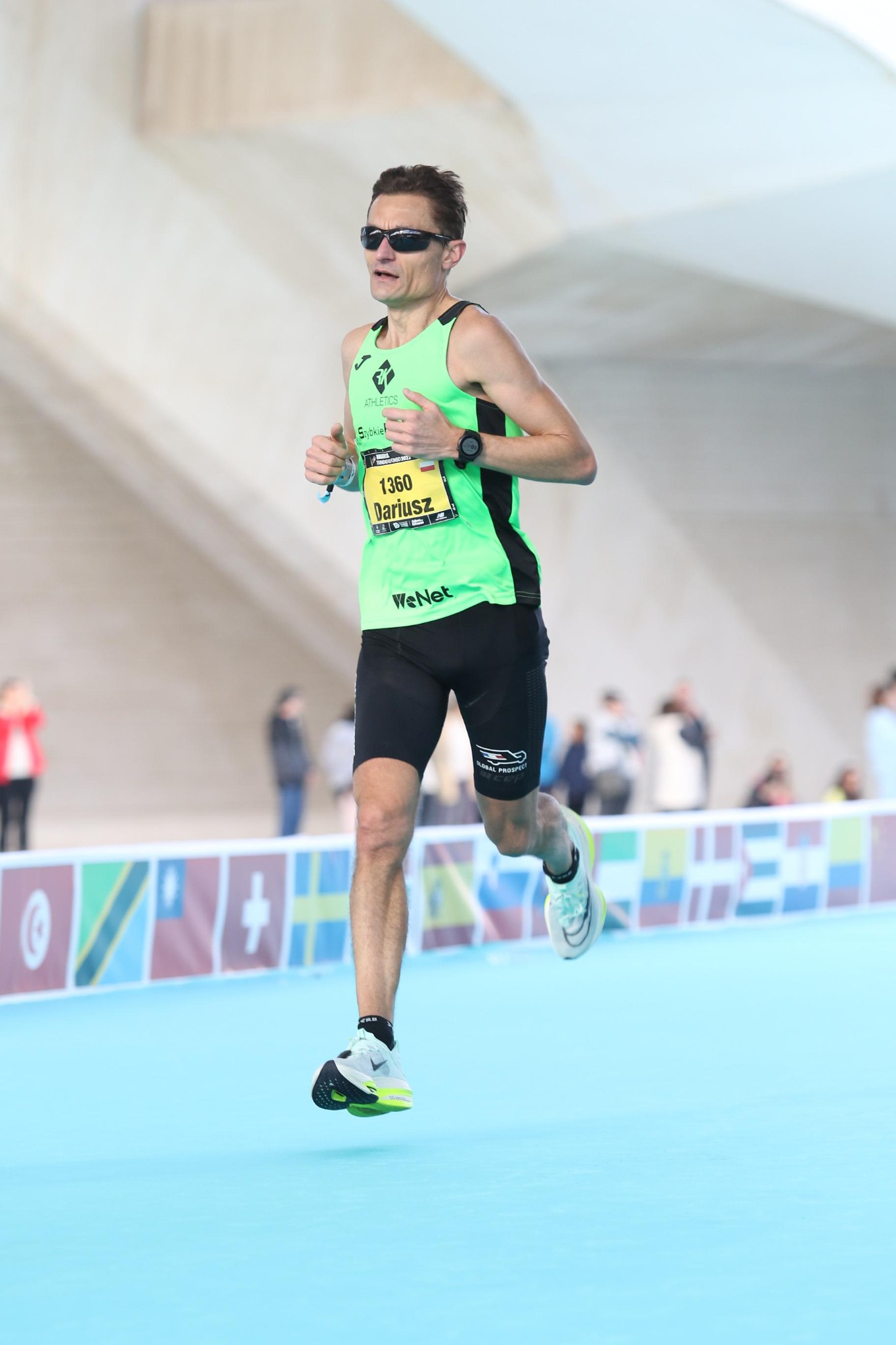 Mężczyzna w okularach przeciwsłonecznych i sportowym stroju biegnie podczas maratonu, widoczny numer startowy 1360, na nadgarstku zegarek sportowy, w tle publiczność i elementy infrastruktury.