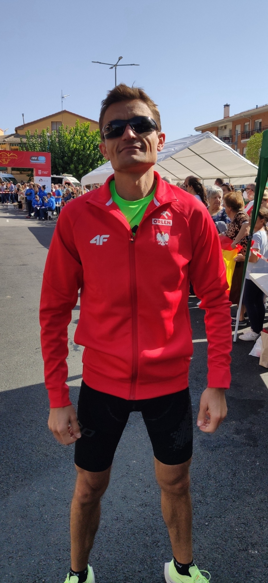 Mężczyzna w czerwonej kurtce sportowej z logo 4F i Orlen, okularach przeciwsłonecznych, na tle tłumu i budynków miejskich, prawdopodobnie po biegu.