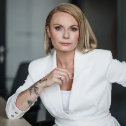 Portret kobiety w białym garniturze, z tatuażem na przedramieniu, siedzącej przy biurku w jasnym pomieszczeniu.