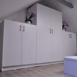 Biała zabudowa szaf wnękowych pod skosem dachu, uzupełniona storczykami w doniczkach, z pufą na pierwszym planie, widok na fragment sypialni.