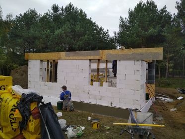 Wznoszenie ścian parterowego domu z białych bloczków, widoczny robotnik w niebieskiej koszulce na kolanach, ocieplenie fundamentów, w tle las i pochmurne niebo.