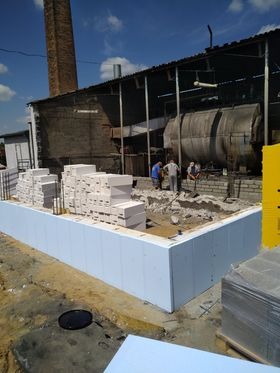 Fundament domu w trakcie budowy: białe bloczki izolacyjne na zewnątrz, palety z białymi bloczkami w tle, robotnicy przy betonowaniu. Widoczny komin i budynek gospodarczy.