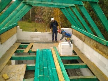 Budowa domu w trakcie: widok z góry na szkielet dachu z zielonych belek, ściany z białych bloczków i ocieplenie z wełny mineralnej. Dwóch pracowników w strojach roboczych układa kolejne bloczki.