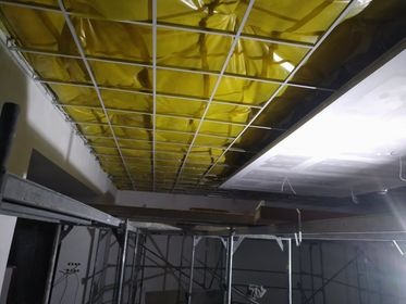 Montaż sufitu podwieszanego z płyt kartonowo-gipsowych, widoczna metalowa konstrukcja rusztu wypełniona żółtą folią izolacyjną, w tle rusztowanie i fragment ściany.
