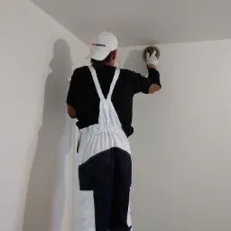 Pracownik w kombinezonie i czapce szlifuje ścianę w rogu pomieszczenia za pomocą szlifierki oscylacyjnej, przygotowując ją do dalszych prac wykończeniowych.