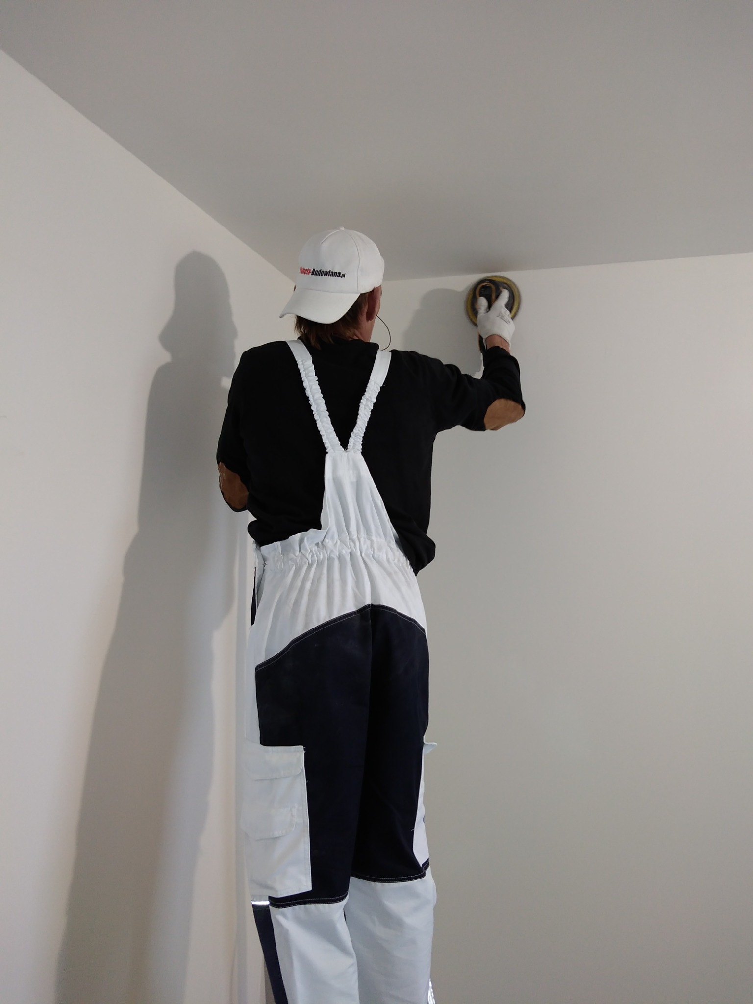 Pracownik w kombinezonie i czapce szlifuje ścianę w rogu pomieszczenia za pomocą szlifierki oscylacyjnej, przygotowując ją do dalszych prac wykończeniowych.