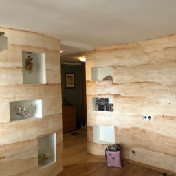 Uslugi Budowlane Piotr Towsciuk - Ściana działowa z dekoracyjną, warstwową fakturą w odcieniach beżu i brązu, z wnękami wypełnionymi dekoracjami, widoczne gniazdka elektryczne i torba na podłodze.