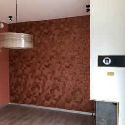 Uslugi Budowlane Piotr Towsciuk - Pokój po wykończeniu z dekoracyjną ścianą w odcieniach brązu z geometrycznym wzorem, drewnianą lampą wiszącą i nowoczesnym kominkiem z zabezpieczeniem.