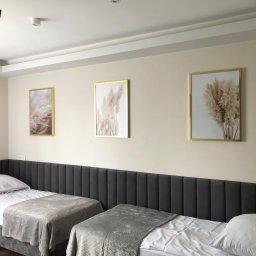 Uslugi Budowlane Piotr Towsciuk - Jasne wnętrze pokoju z dwoma łóżkami pojedynczymi, szarym wezgłowiem i trzema obrazami w złotych ramach przedstawiającymi pampasową trawę na beżowej ścianie.