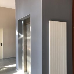 Uslugi Budowlane Piotr Towsciuk - Wnętrze klatki schodowej z windą, ściany pomalowane w odcieniach szarości, widoczny grzejnik panelowy i fragment podłogi z płytek, wpadające promienie słoneczne.