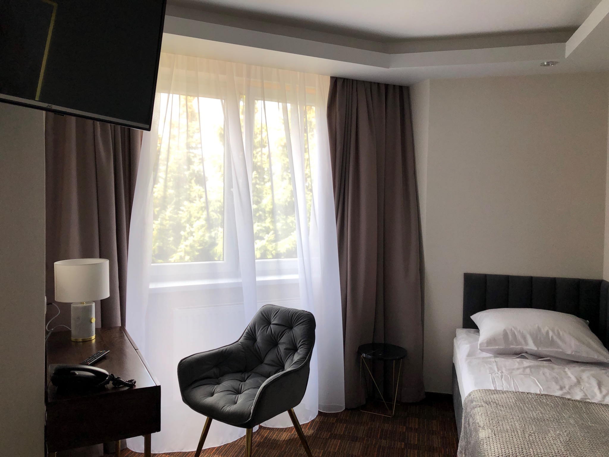 Wnętrze pokoju hotelowego po odświeżeniu, z ciemnymi zasłonami, biurkiem z lampką nocną i tapicerowanym krzesłem na złotych nogach, widok na łóżko i okno.