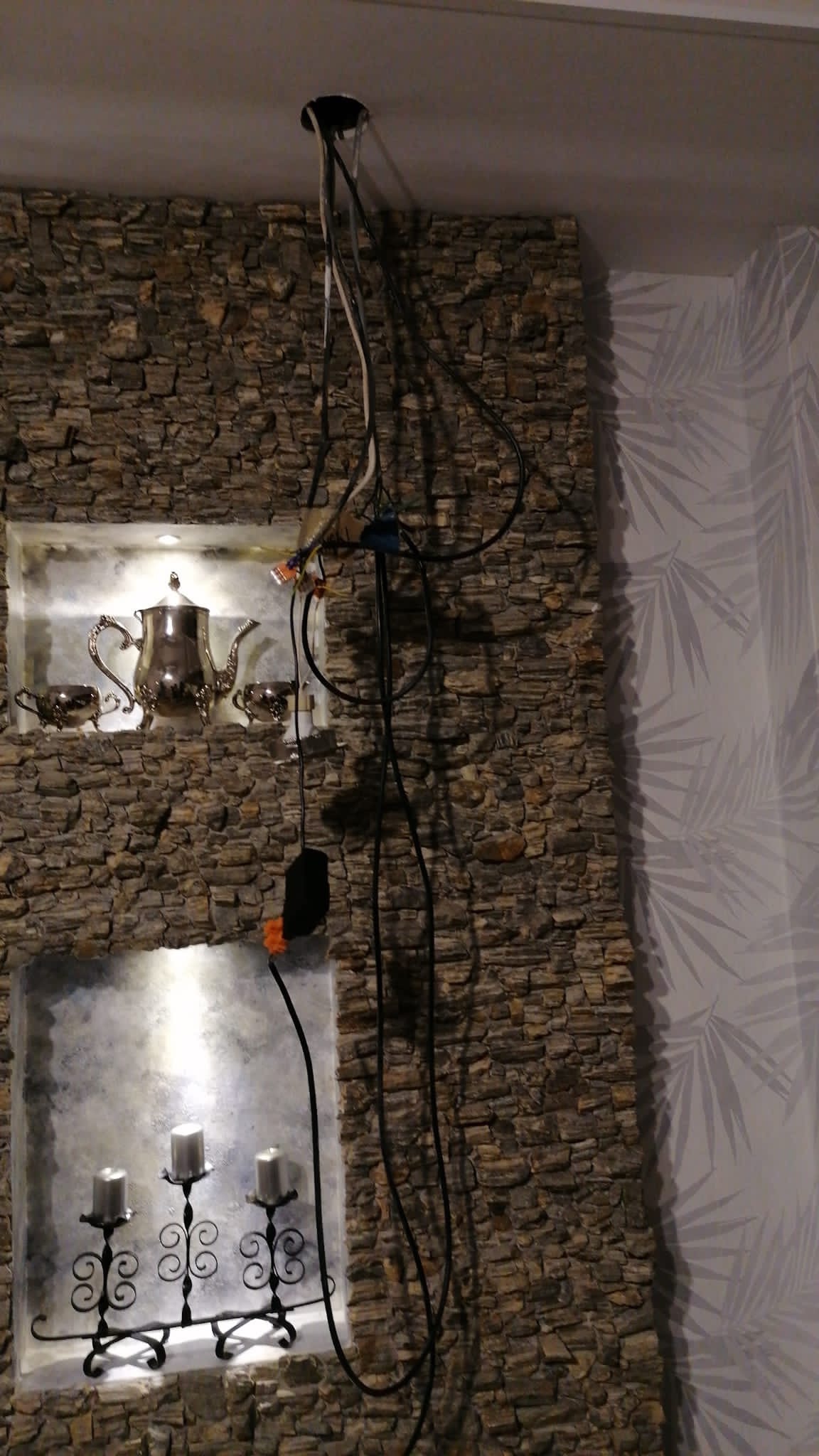 Niedokończone podłączenie lampy sufitowej z widocznymi kablami na tle ściany z kamienia dekoracyjnego z wnękami, w których znajdują się srebrna zastawa i świeczniki.
