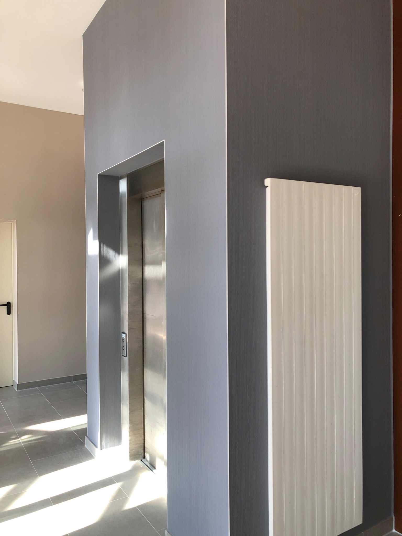 Wnętrze klatki schodowej z windą, ściany pomalowane w odcieniach szarości, widoczny grzejnik panelowy i fragment podłogi z płytek, wpadające promienie słoneczne.