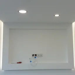 Biała ściana z wnęką pod telewizor, oświetlenie LED w suficie i wnęce, gniazdka elektryczne i kabel antenowy, śrubokręt leżący na półce.