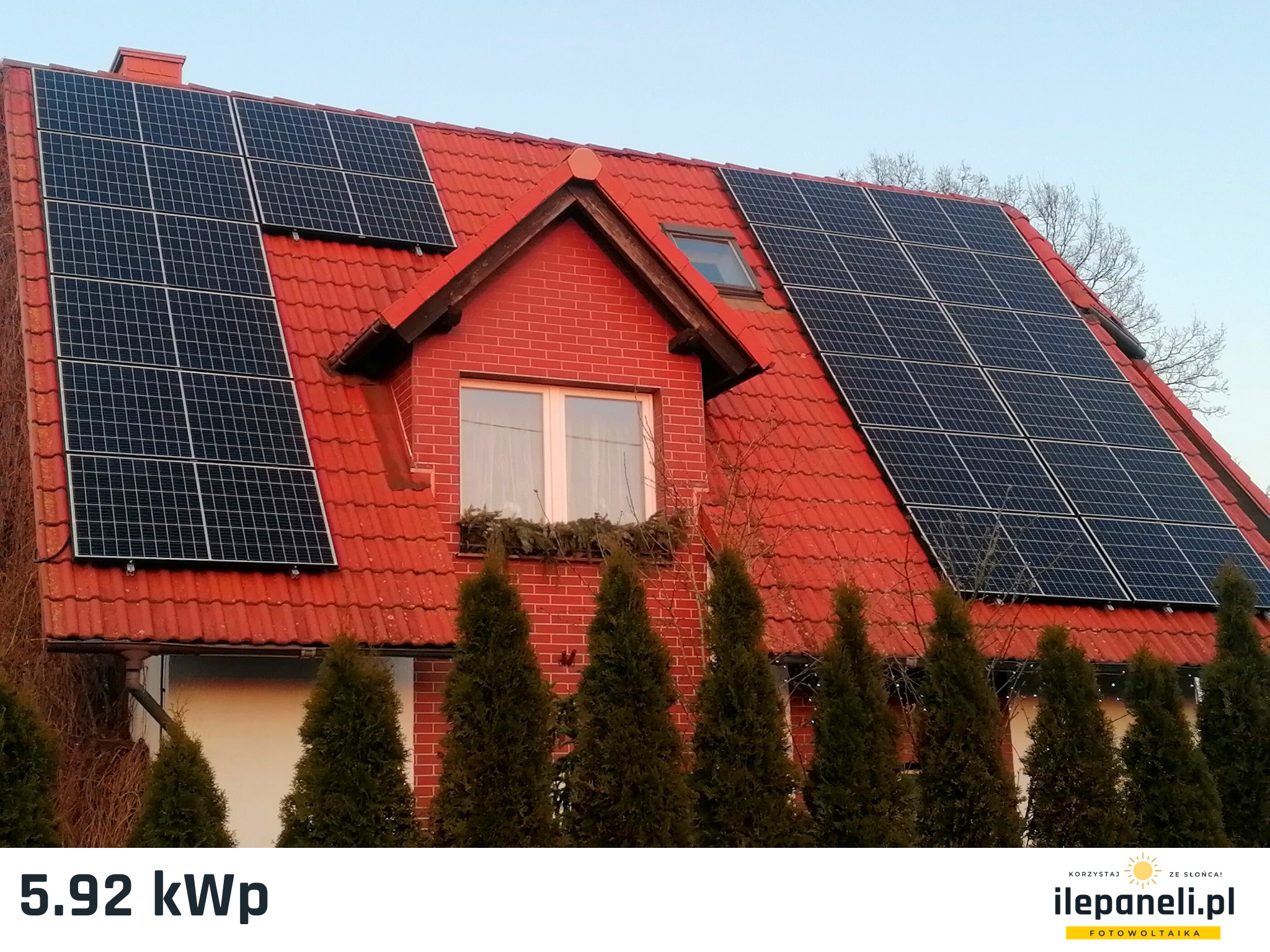 Czerwony dach domu jednorodzinnego z zamontowanymi panelami słonecznymi, widoczne również iglaki w ogrodzie.
