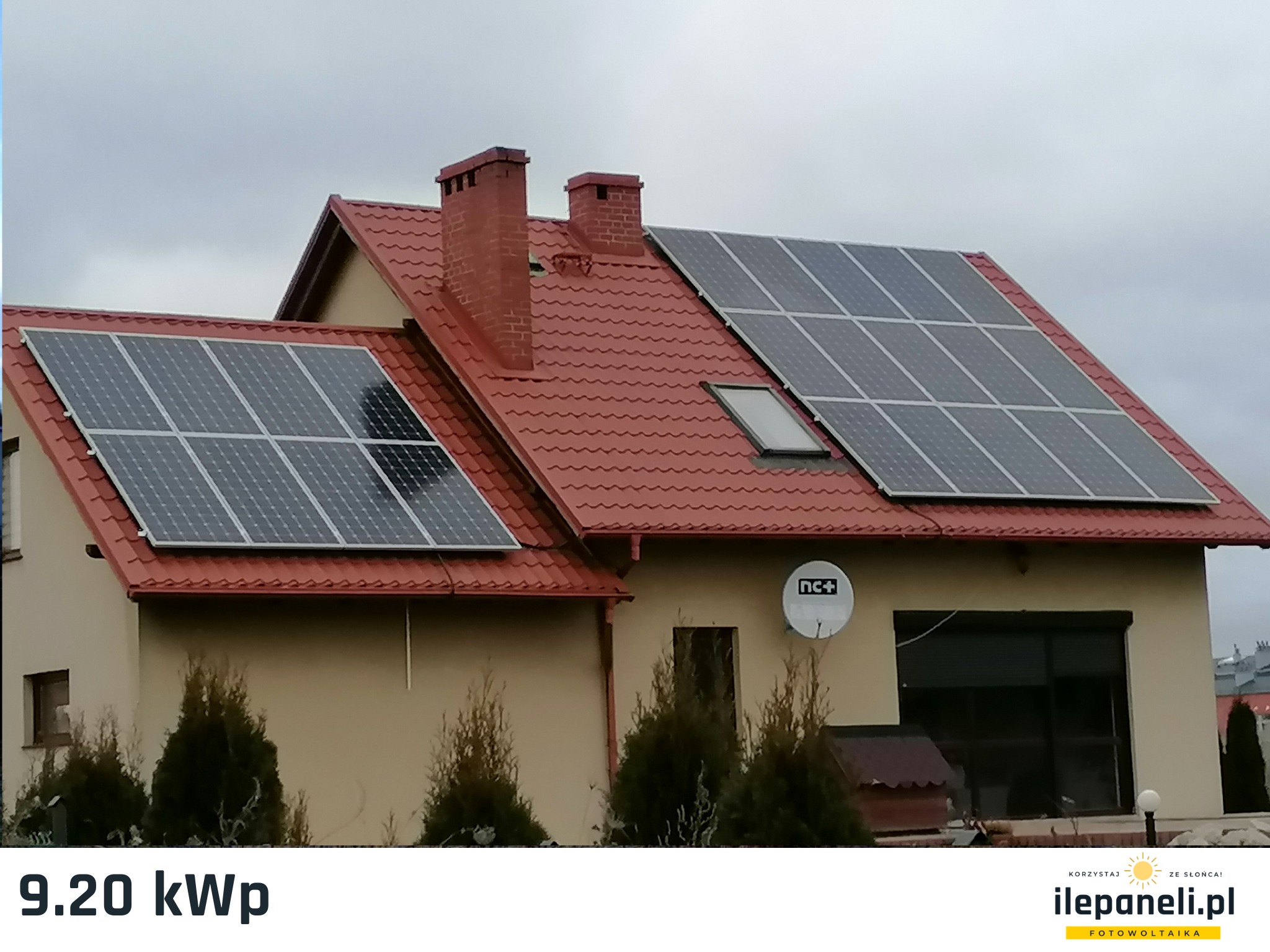 Dom jednorodzinny z panelami słonecznymi na czerwonym dachu, widok z dołu w pochmurny dzień, antena satelitarna na ścianie.