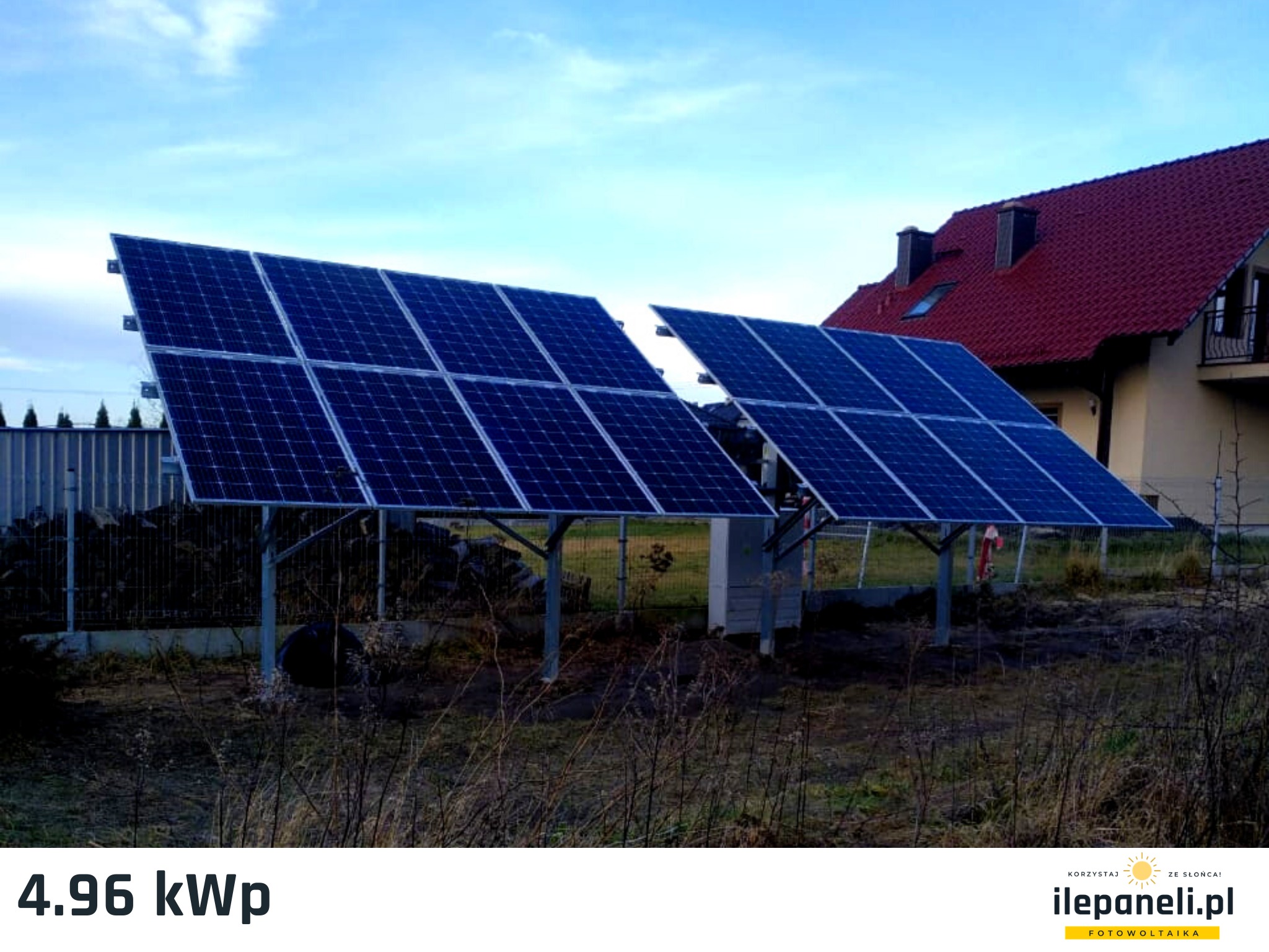 Dwa panele fotowoltaiczne o mocy 4.96 kW na metalowych konstrukcjach, zamontowane na gruncie przed domem z czerwoną dachówką.