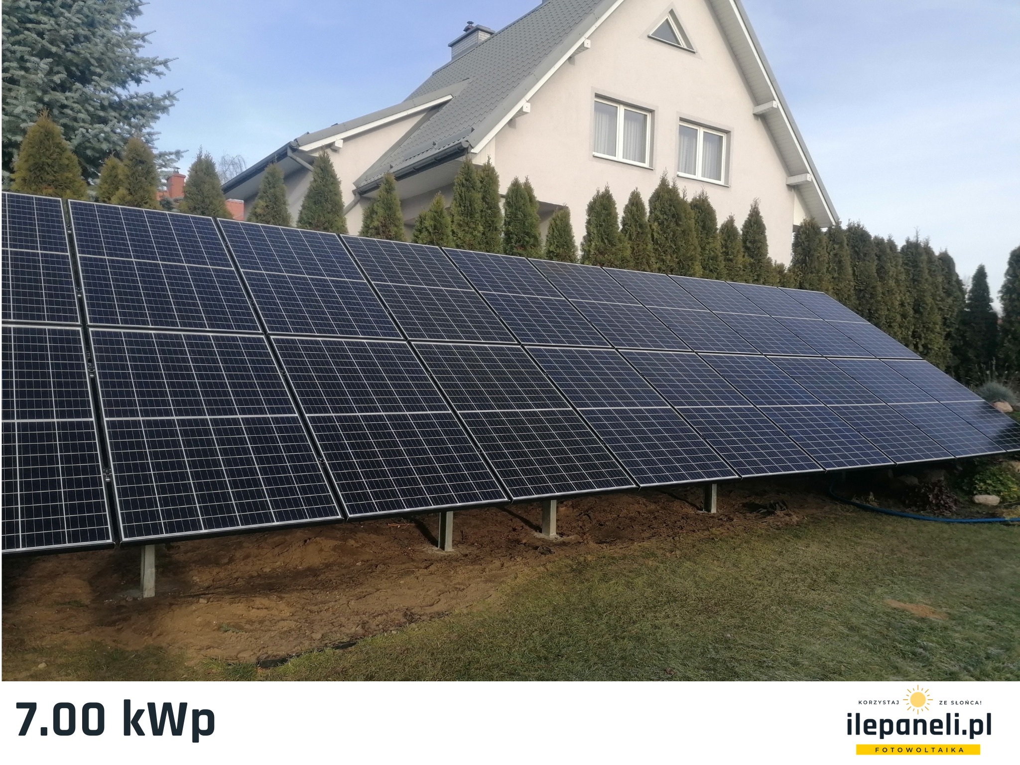 Panel słoneczny o mocy 7.00 kWp zamontowany na ziemi przed domem jednorodzinnym z szarym dachem i rzędem iglaków w tle.