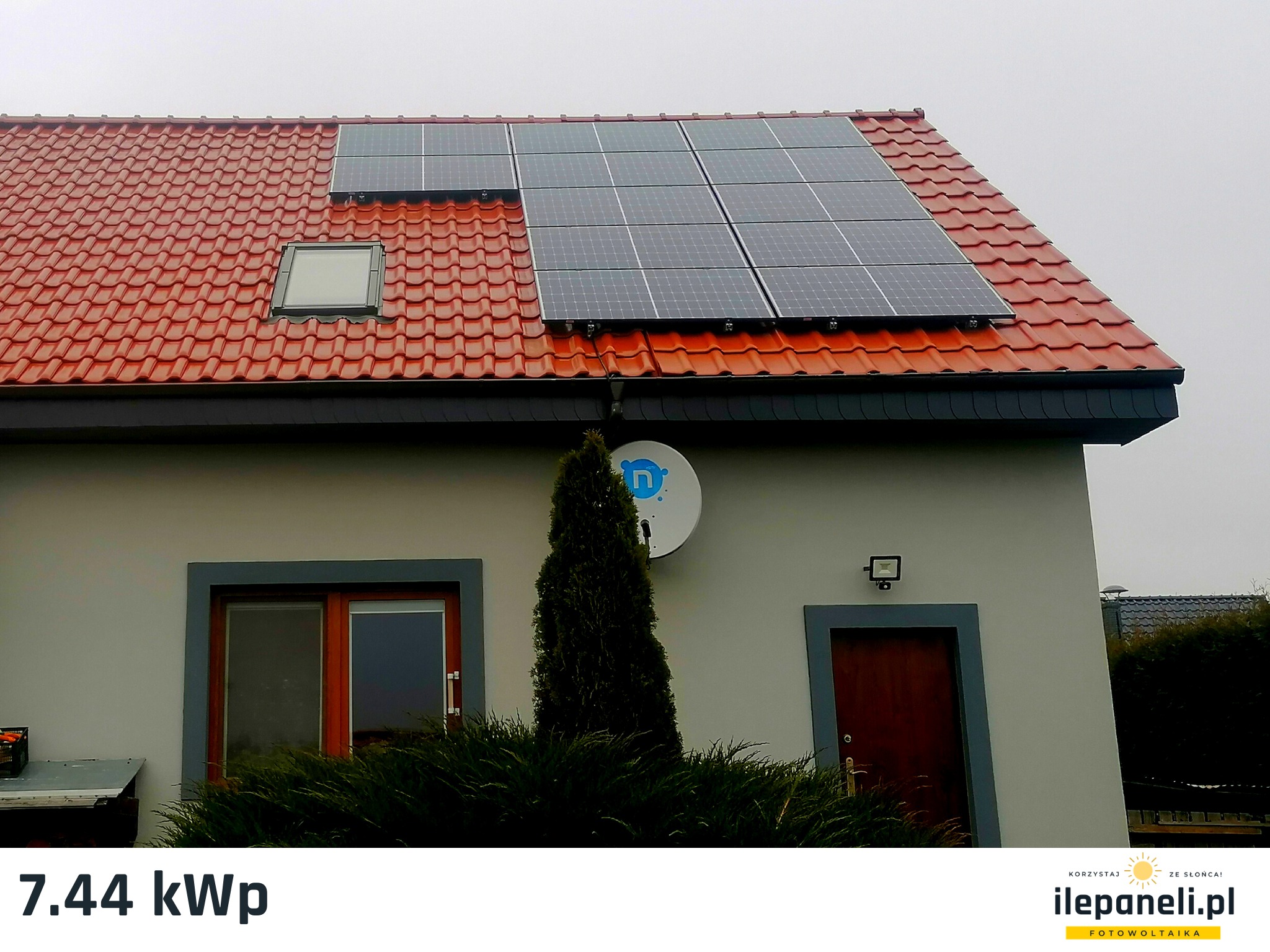 Instalacja fotowoltaiczna na dachu domu z czerwoną dachówką, widoczny satelitarny talerz antenowy i krzew przed budynkiem. Część paneli ułożona nad oknem dachowym.