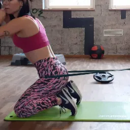 Kobieta w różowym topie i wzorzystych legginsach klęczy na macie Reebok, wykonując ćwiczenie z gumą oporową przymocowaną do żółtych słupków w siłowni.