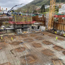 Szeroki kadr na plac budowy z widocznymi zbrojeniami fundamentów, betonową konstrukcją w szalunkach, pracownikami w odblaskowych kamizelkach i żurawiem do betonu w tle, z zabudową miejską...