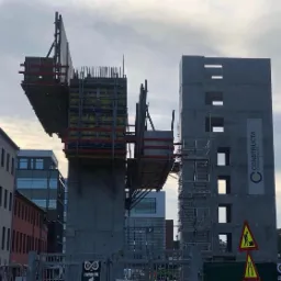 Konstrukcja żelbetowa w trakcie budowy w Gorlicach, widoczne szalunki, zbrojenie oraz fragment gotowego budynku z rusztowaniem i banerem reklamowym.