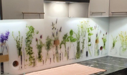 Szklany panel kuchenny z motywem ziół i kwiatów w przezroczystych wazonach, widoczne gniazdka elektryczne w panelu.