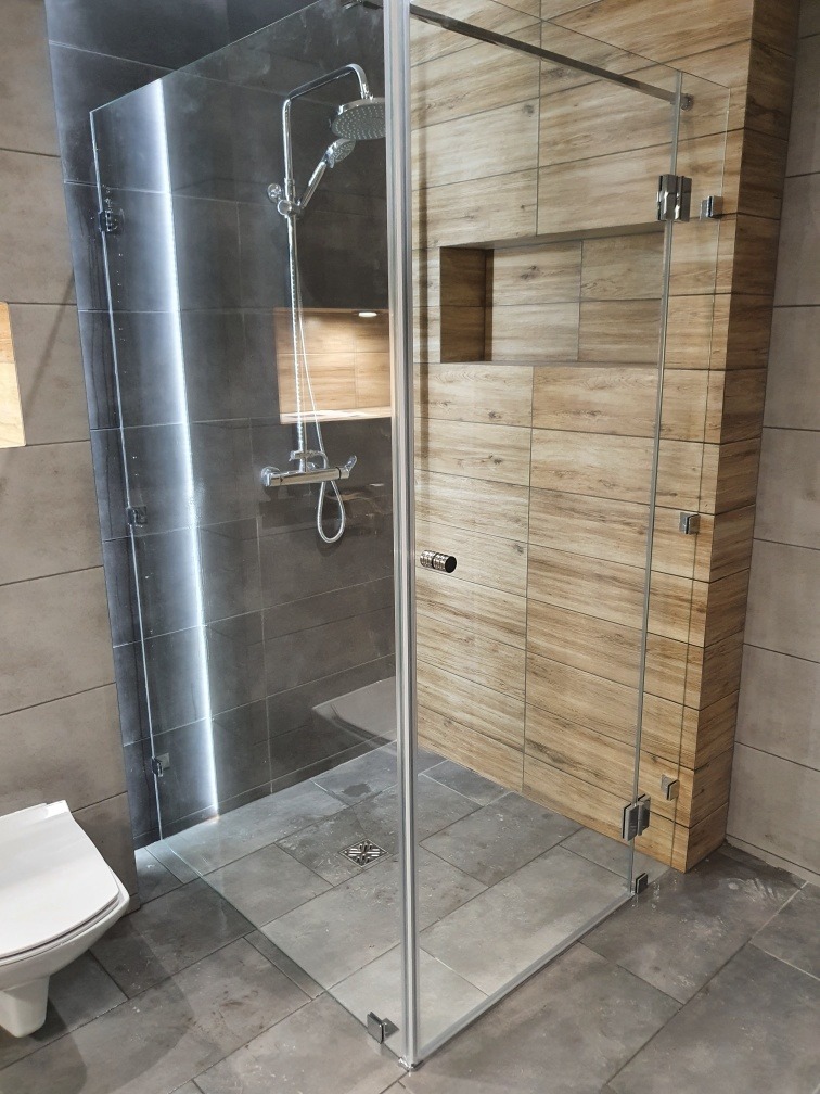 Kabina prysznicowa ze szklaną ścianą i drzwiami, z chromowaną armaturą, widok na drewnopodobne płytki ścienne z wnęką, szare płytki podłogowe i toaletę w tle.